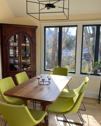 Dining room with wooden table, lime-green chairs, large windows overlooking trees, and dark wood cabinet.