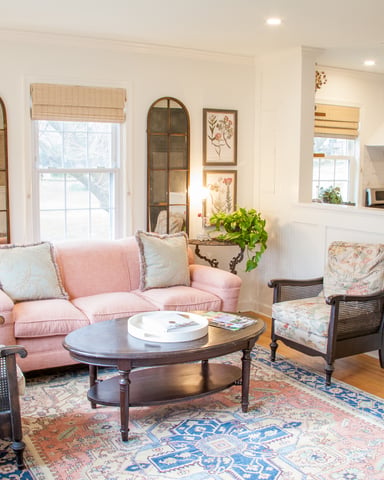 Bright living room with pink sofa, dark wood coffee table, botanical artwork, and open kitchen visible in background