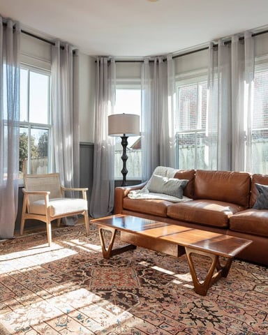 Bright living room with cognac leather sofa, mid-century modern furniture, patterned rug, and floor-to-ceiling windows with sheer curtains overlooking brick