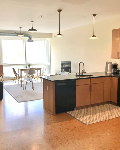 Open-plan kitchen and living area with wood cabinets, black appliances, island counter, and seating area with windows.