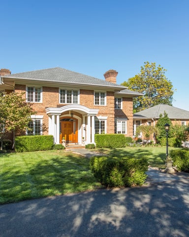 Two-story brick colonial house with white portico entrance, manicured lawn, and mature trees surrounding property