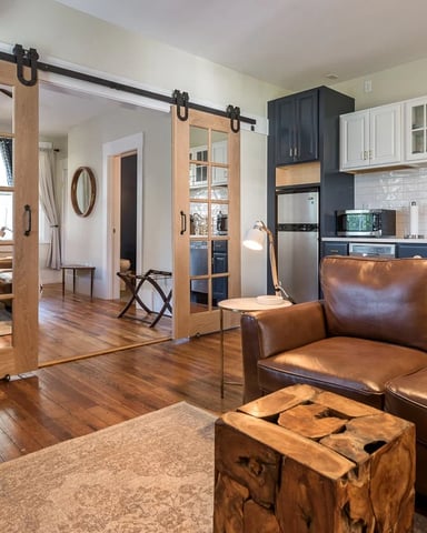 Open-plan living room and kitchen with brown leather sofa, barn doors, wood floors, and stainless steel appliances.