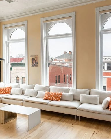 Bright living room with cream sectional sofa, orange accent pillows, and large arched windows overlooking brick buildings