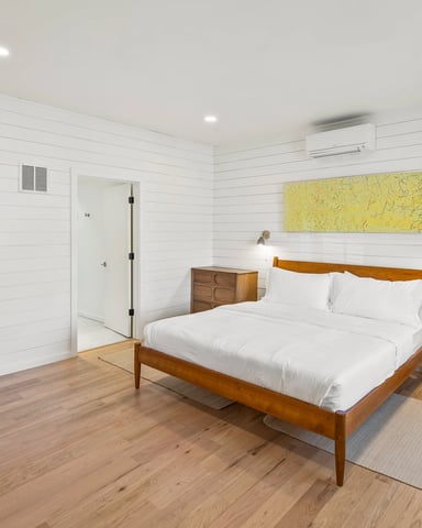 Bright bedroom with white shiplap walls, wooden bed frame, white bedding, wood nightstands, and colorful artwork on walls