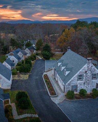 Aerial view of stone and white houses with dark roofs on a curved driveway, surrounded by autumn trees and distant mountains at sunset.