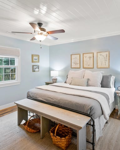 Light blue bedroom with queen bed, wooden nightstands with lamps, framed botanical prints, ceiling fan, window with forest view, and wooden bench at foot of