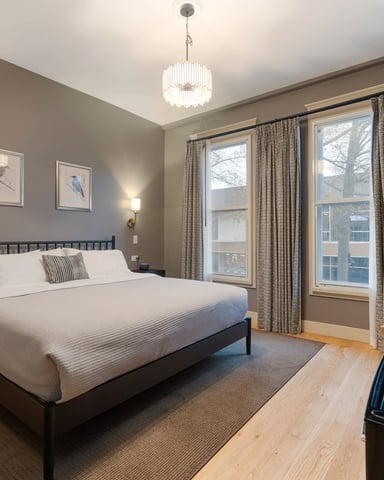 Modern bedroom with queen bed, taupe walls, large windows with curtains, pendant light, and city view