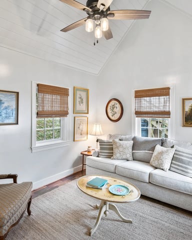 Bright living room with white walls, vaulted ceiling, ceiling fan, gray sofa, upholstered armchair, landscape paintings, and windows with woven shades.