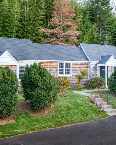 Single-story cottage with cedar shingle and white trim, slate roof, surrounded by evergreen shrubs and forest backdrop.