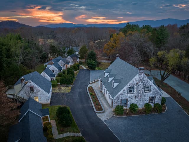 Aerial view of stone houses with slate roofs on a curved driveway, surrounded by autumn trees and distant mountains at sunset.